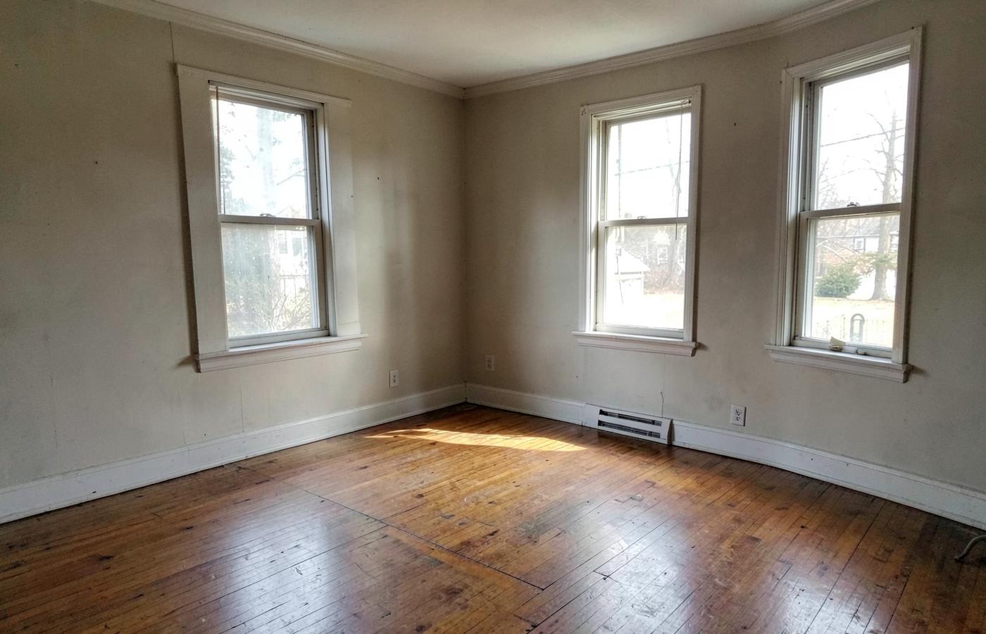 Empty living room with hardwood floors and bare walls lit by three tall windows